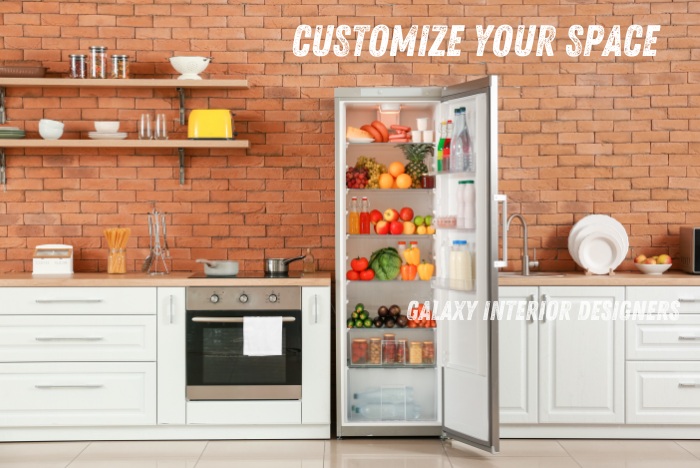 Galaxy Interior Designers in Chennai specialize in creating personalized kitchen spaces. This image showcases a modern modular kitchen with white cabinetry, open wooden shelves, a stainless steel oven, and a fully stocked refrigerator filled with fresh fruits and vegetables